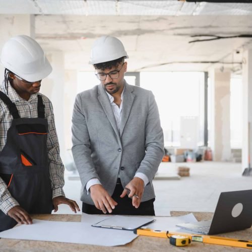 Architect and builder wearing hardhats discussing blueprint on table with laptop and spirit level in building under construction