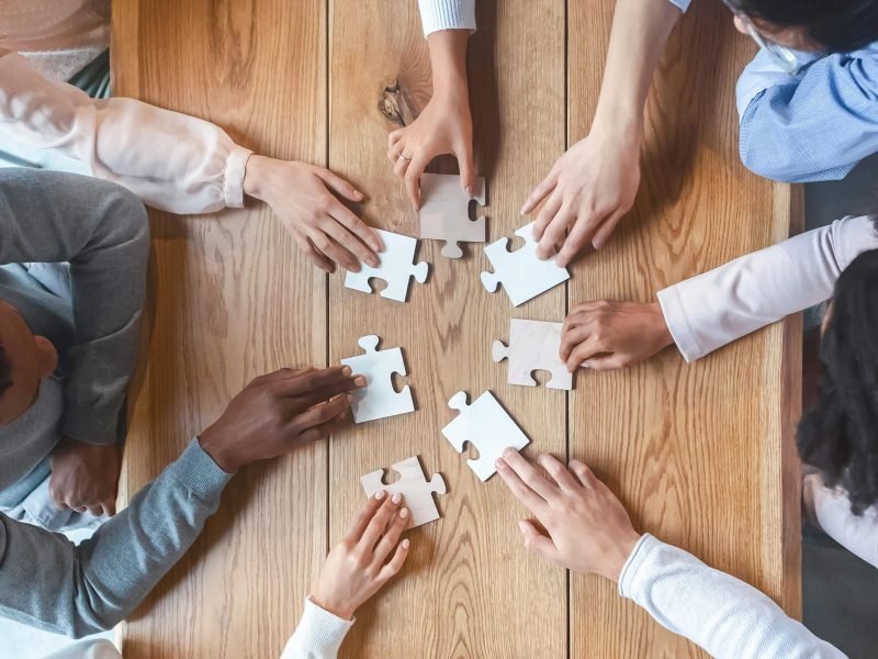 Collaboration concept, top view of creative multiracial team sitting around table and putting together puzzle pieces, cropped